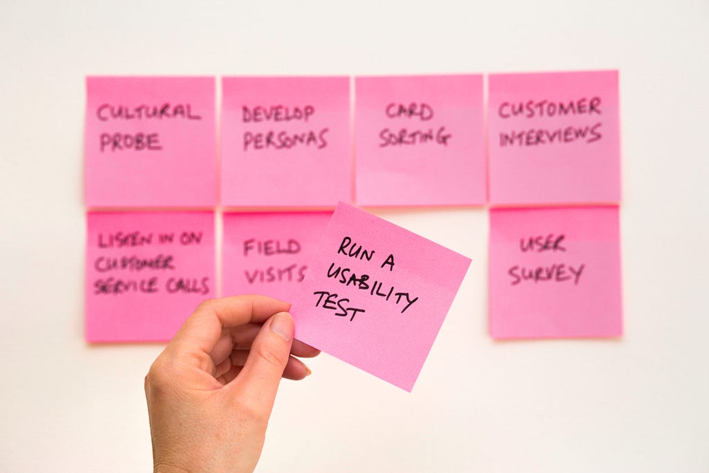 Sticky notes named with UX research methods. One sticky note is held by a hand in the foreground named “Run a Usability Test”