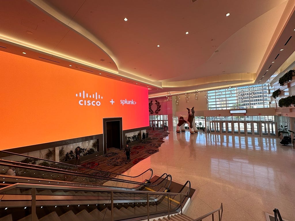 A large, modern indoor space with a grand staircase, a massive digital screen displaying Cisco + Splunk, and tall windows providing extensive natural light.