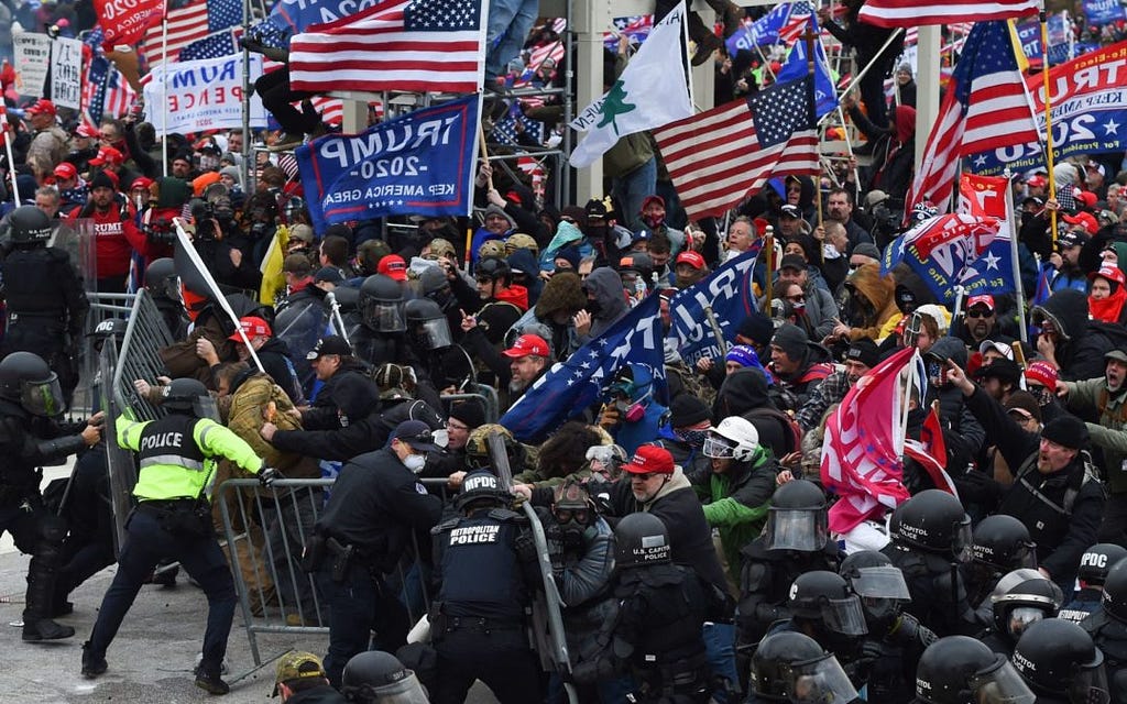 A photo of the January 6th insurrection that depicts the rioters pushing through a security barrier guarded by the police while carrying american and MAGA flags.