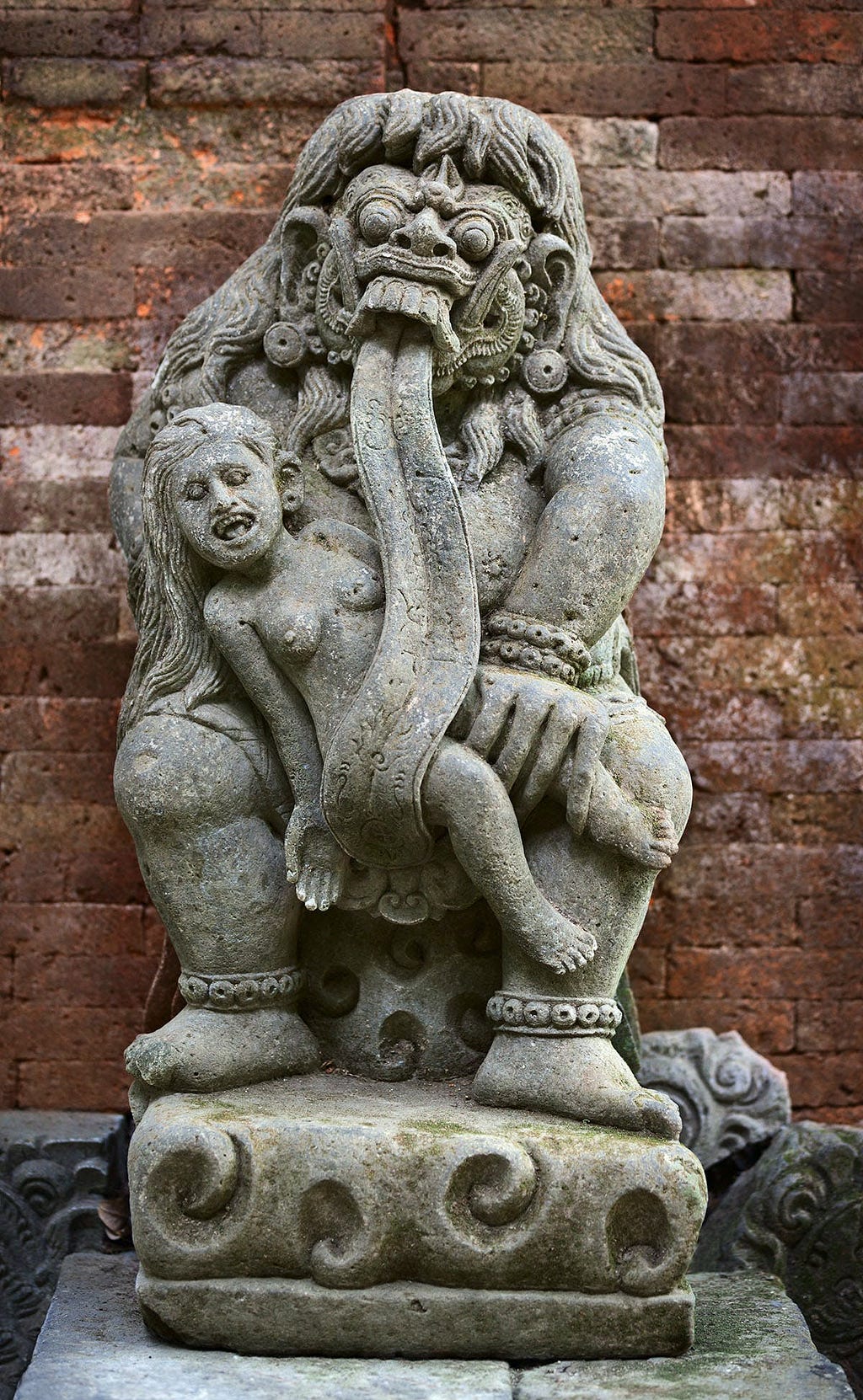 Statue of Rangda, the mythical Queen of Leyak in Balinese folklore, depicted with fierce facial expressions, a long protruding tongue, and a small figure clutched in her left arm. The statue shows intricate carvings and stands against a backdrop of weathered red bricks.