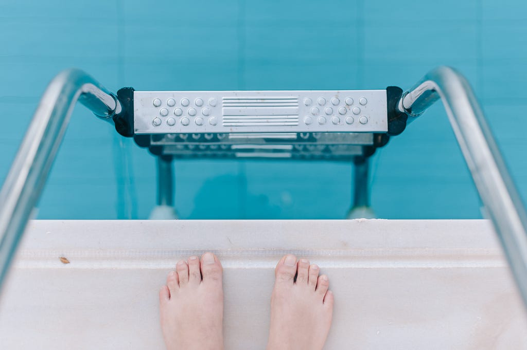 A first person view of someone looking down a poolside ladder with their feet on the pool's edge.