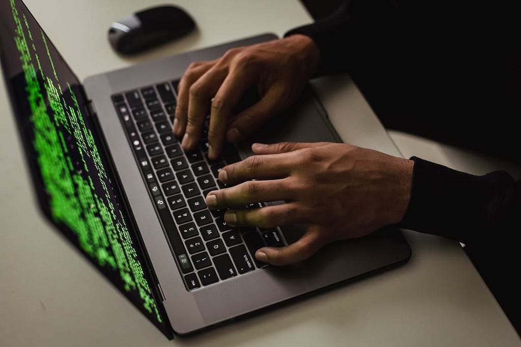 An open laptop computer, viewed from above and to the left. A person’s hands are typing on they keyboard. Blurred green text, that looks like computer code, is displayed on the laptop’s screen in front of a black background.