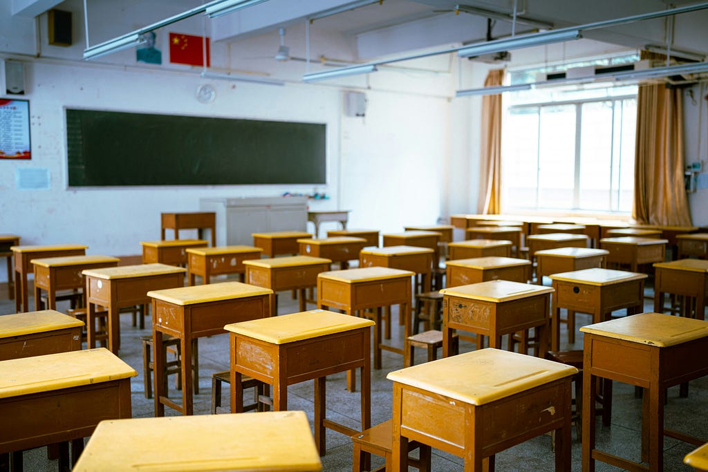 An empty classroom filled with several rows of wooden desks and stools. Lit by sunshine through the window. A blackboard covers much of the front wall.