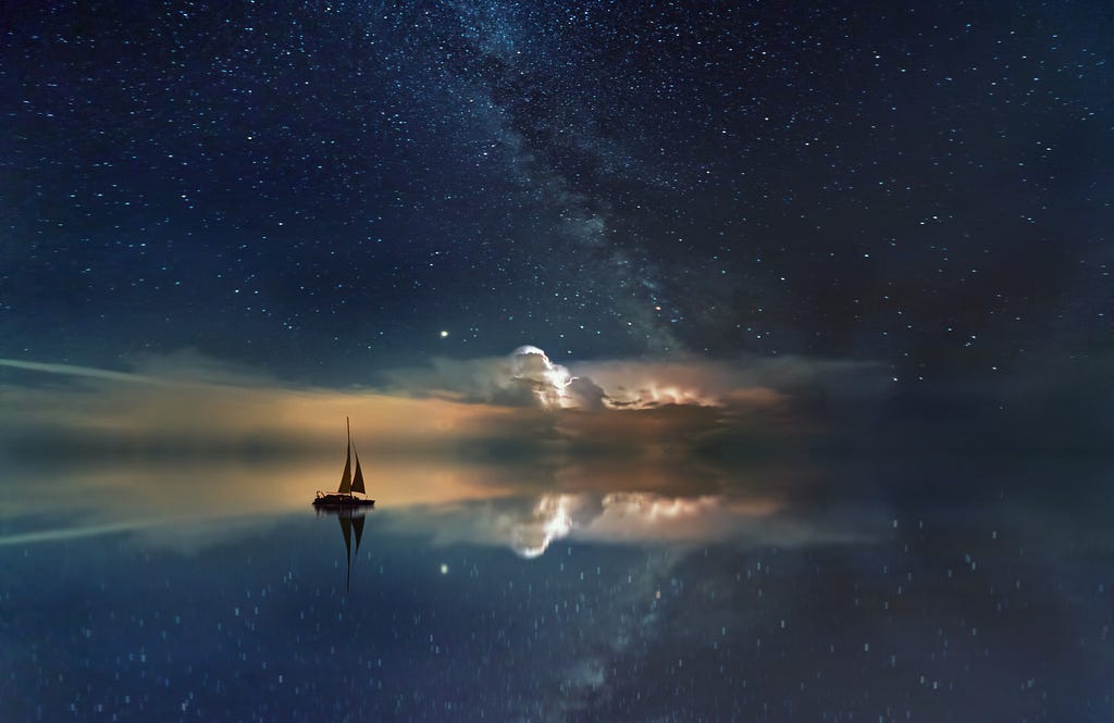 A small boat sails through a tranquil body of water reflecting the starry sky and the sunrise at dawn.