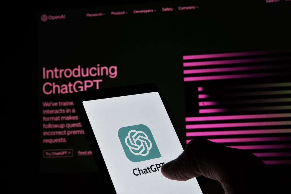 A computer screen showing ChatGPT in the background as well as a mobile device being held with ChatGPT logo on it.