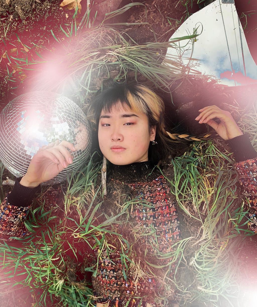 Alice is laying centered on a bed of soil, wrapped in knots of grass, surrounded by a disco ball to her left and a shard of glass to her top right reflecting the blue cloudy sky. She presents as light-skinned East Asian with black and blonde-dyed hair in braids. She holds a willful expression, with hands resting on the ground on either side of her face and reddish-white light swirling around her bust as if in a moment of magic.