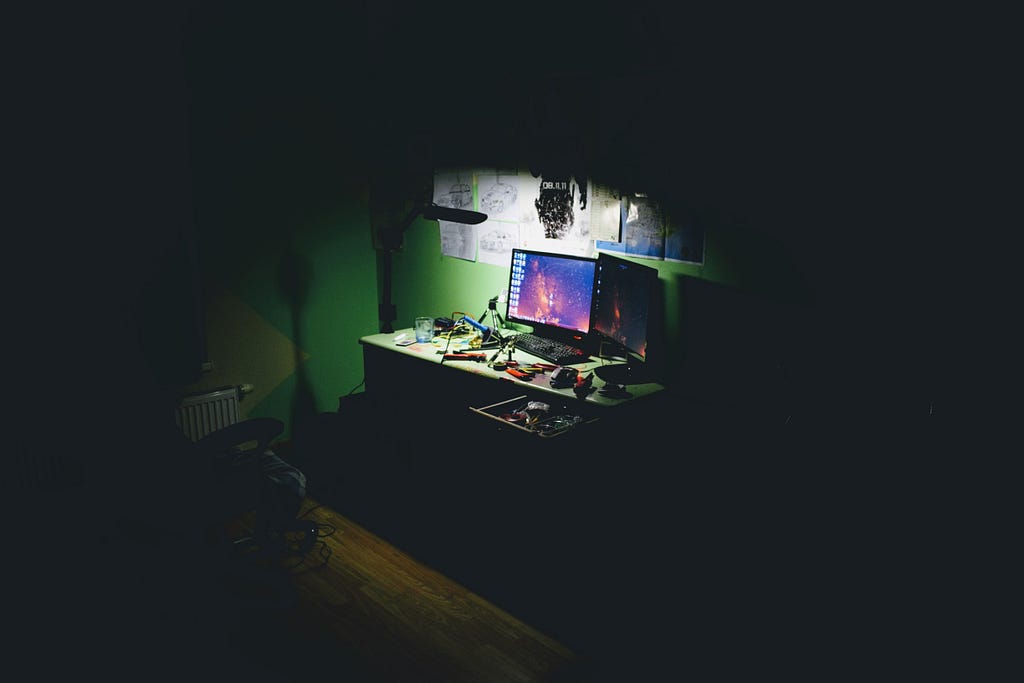 A computer sits in a dark room, dimly lit by a weak light overhead