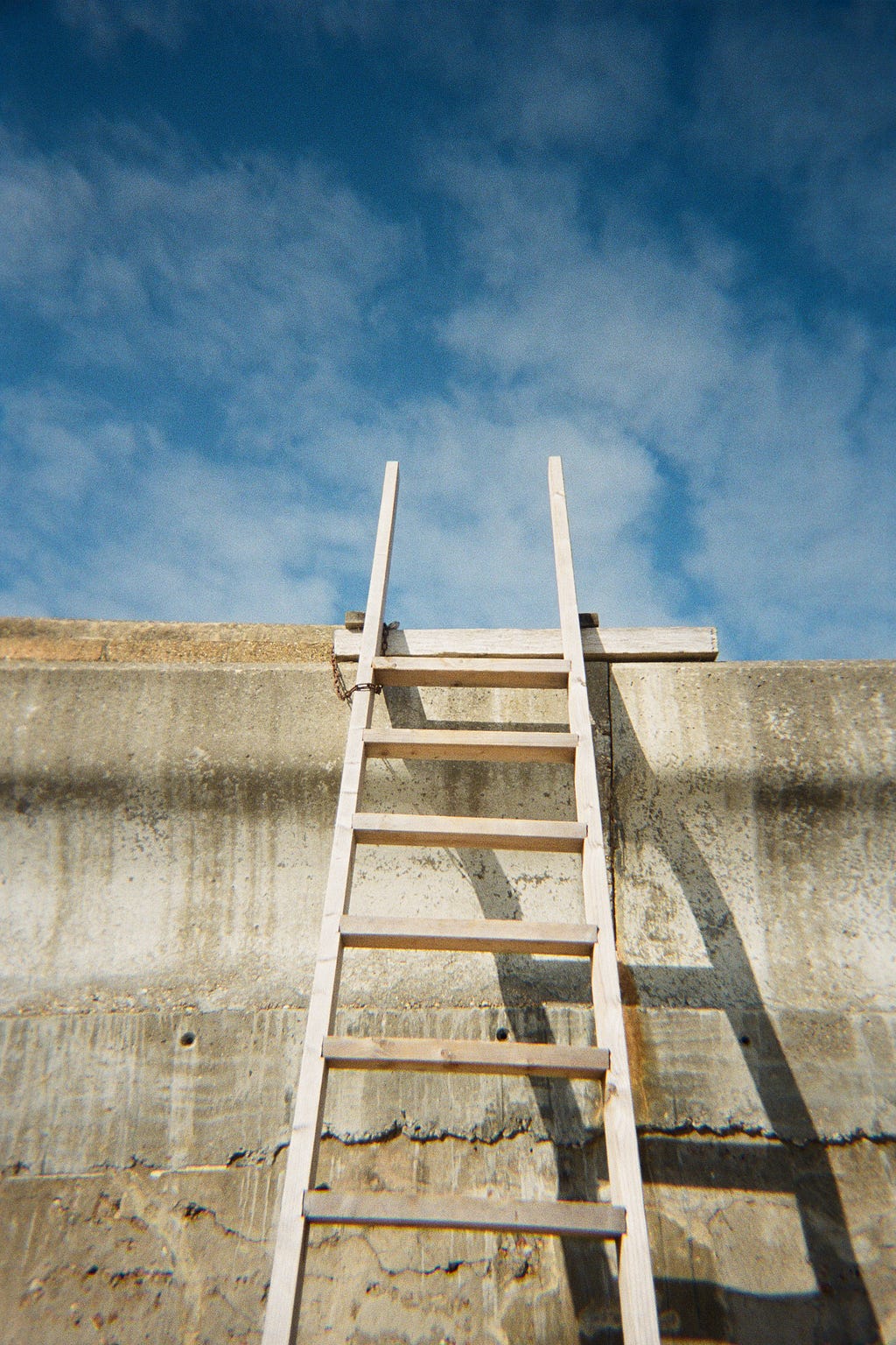 A ladder chained to a concrete wall with a blue sky with puffy white clouds above it