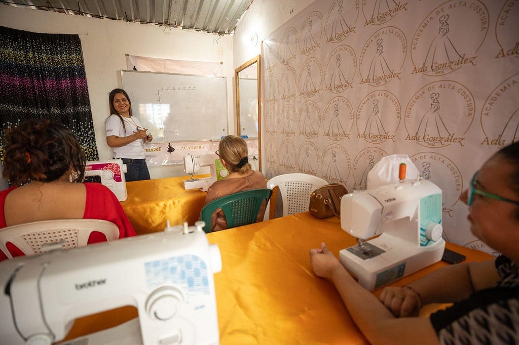 Una mujer da instrucciones en la pizarra a una sala con mujeres sentadas frente a máquinas de coser.