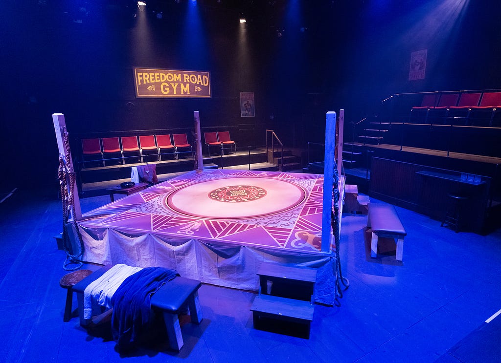 A boxing ring inside a theater, with seats surrounding it and a sign reading “Freedom Road Gym.”