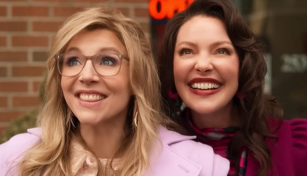 Tully and Kate, on their young adult versions, are shown in a close-up, both widely smiling. Kate is blond and is wearing her big glasses and a pink jacket. Tully has brown, wave hair and is wearing a dark red jacket. She also wears a grey, black and fucsia, thin handkerchief.