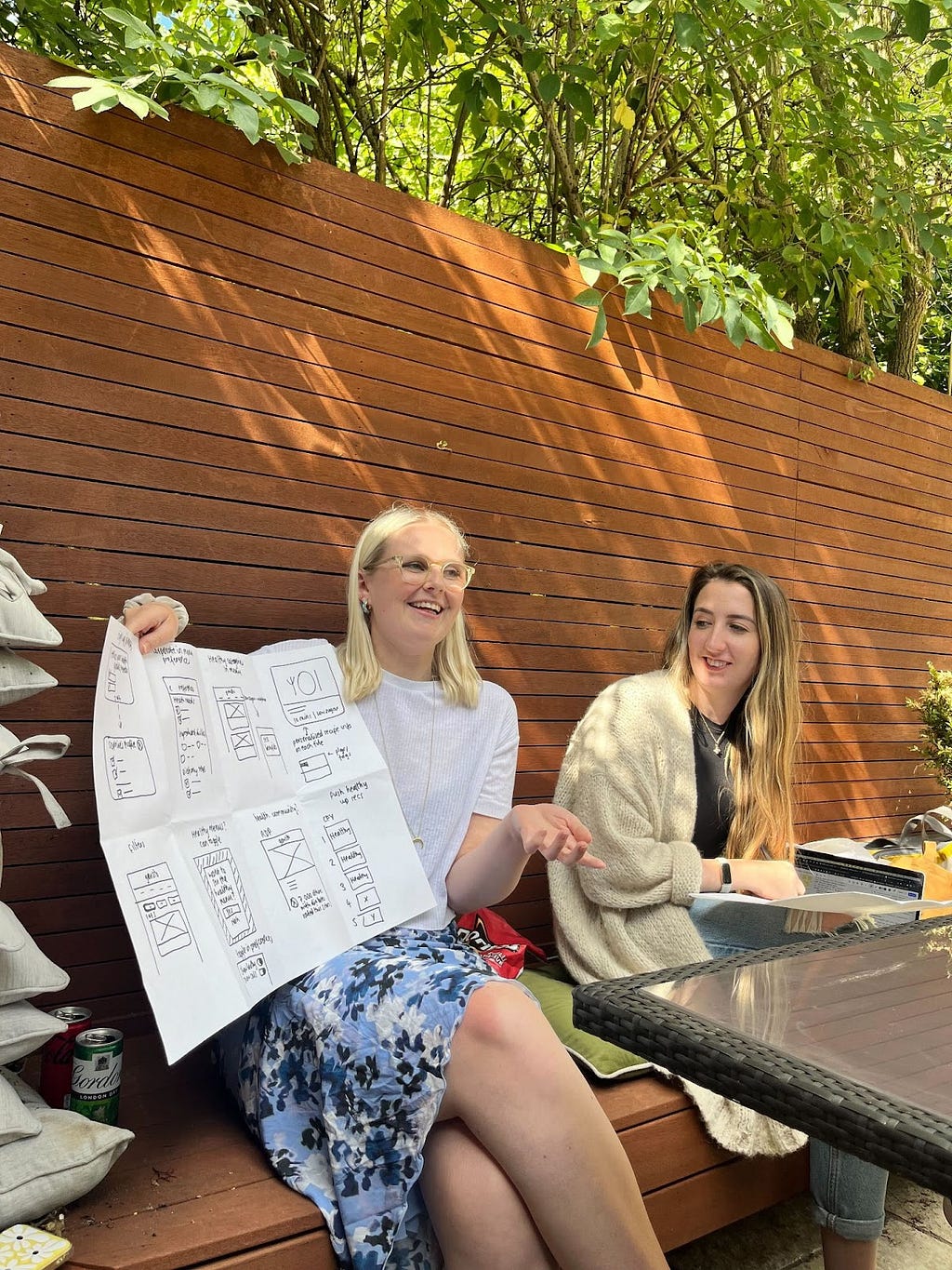 Lady with blonde hair holding up a piece of paper and describing what she’s written on it, next to a lady with darker hair