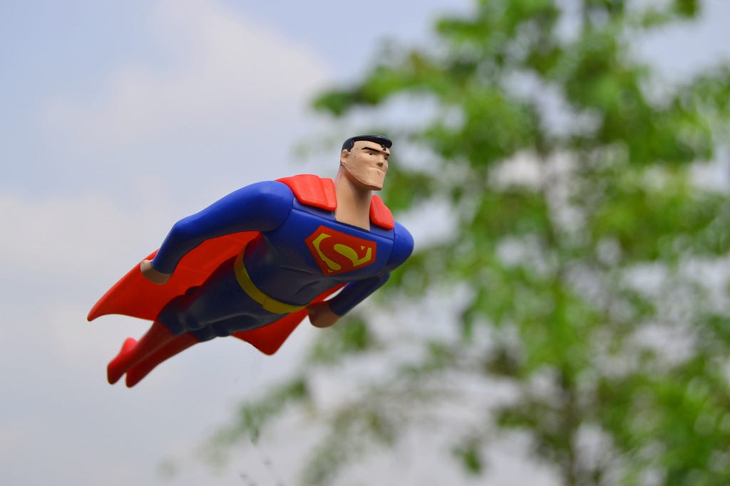 An action figure of Superman flies through the air in front of a green tree