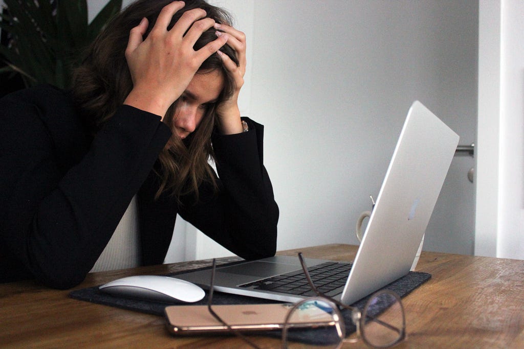A woman gripping her head with her hands, staring at her laptop in an work from home office.