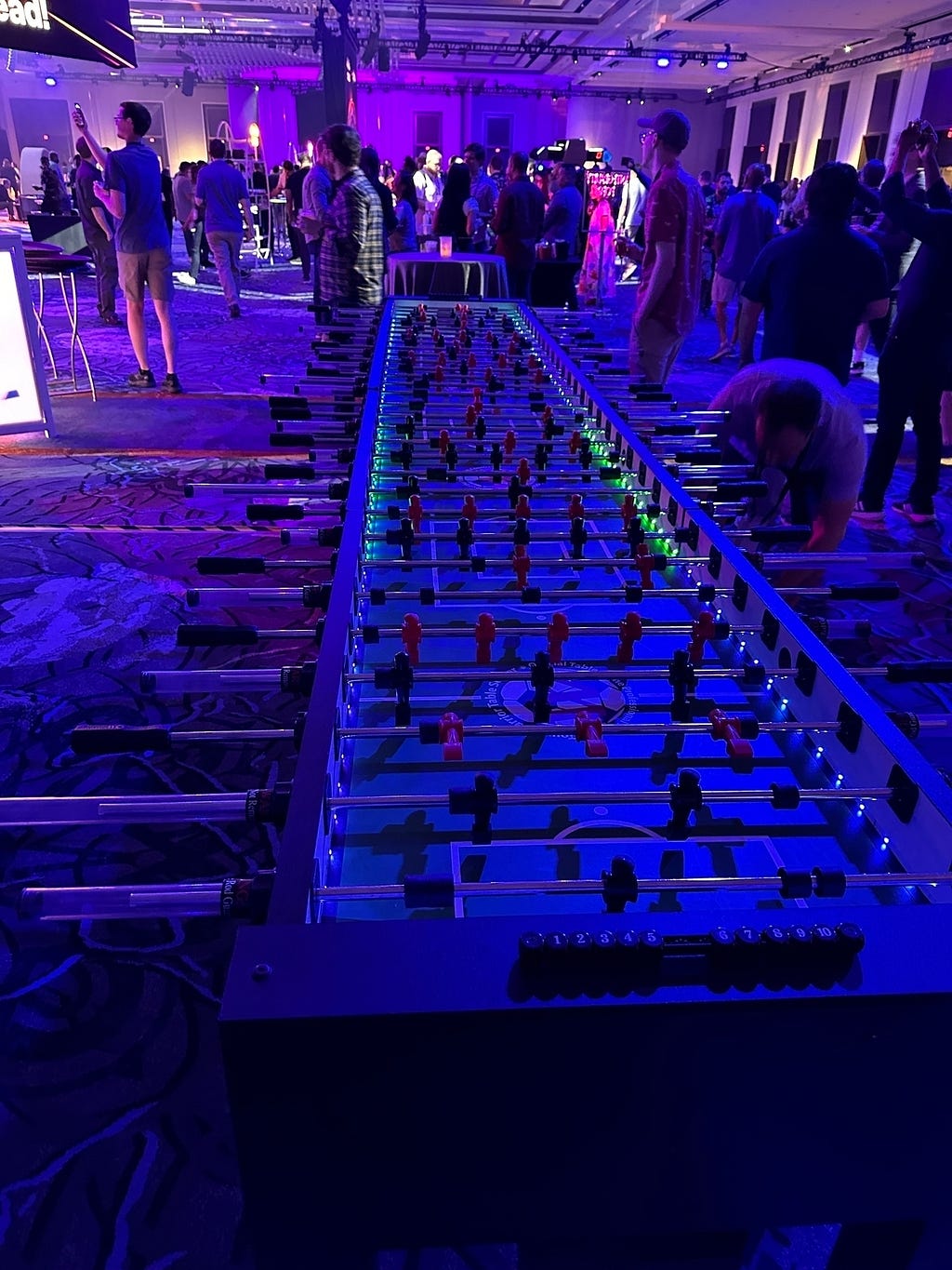 A large room is filled with people socializing and playing a long, multi-player foosball table under vibrant purple and blue lighting.
