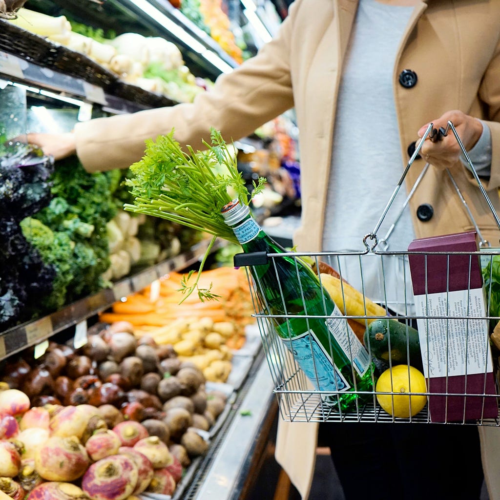 The torso and arm of a white woman wearing a beige coat and grey top as she stands in a supermarket vegetable aisle, holding a basket full of shopping and taking food off the shelves