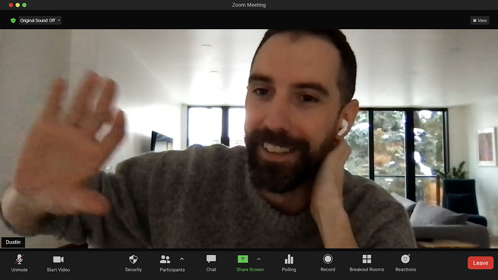 Screenshot from a Zoom call showing Dustin Duffy sitting in a living room wearing a gray sweater, waving and smiling while he talks to the camera.