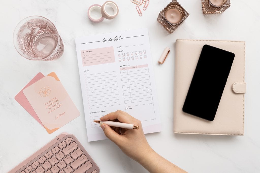 A  flat lay of a to-do list being written on, surrounded by a pink note book with a phone resting on top, some pink stationary, candles and a glass of water.