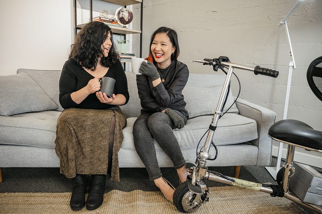 A Latinx disabled woman & an Asian disabled woman smiling at each other, holding mugs. A mobility scooter is to the side.