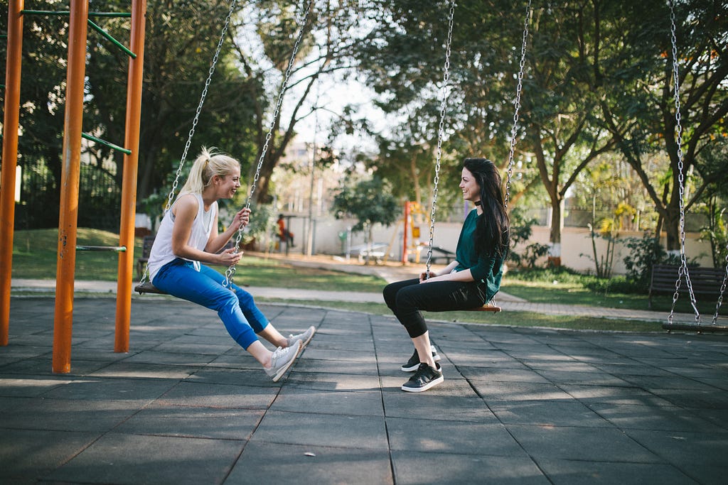 two women, sitting sideways on swings in a park, looking at each other talking and smiling