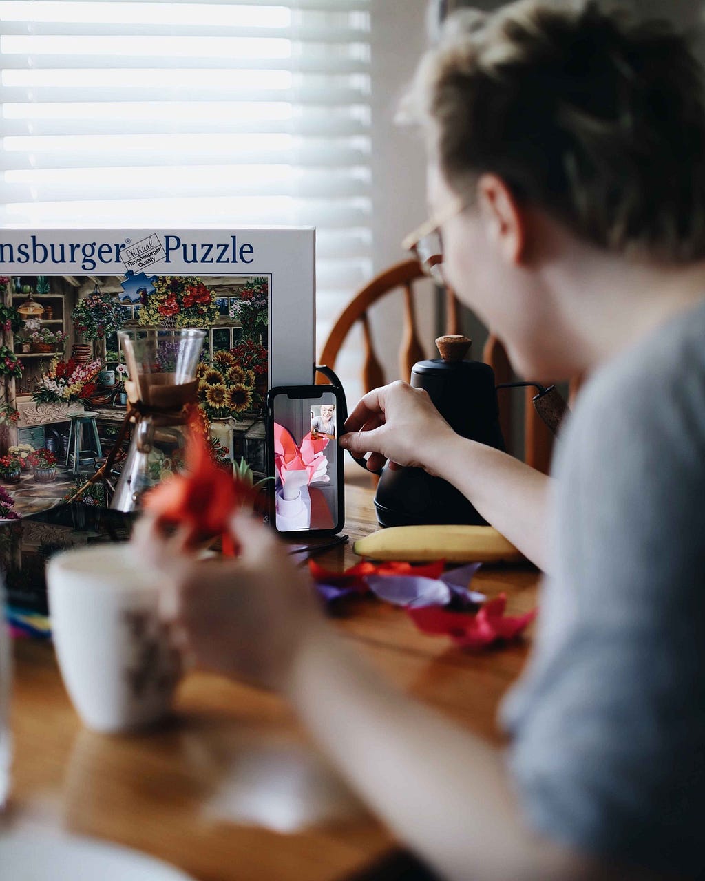 A young person sits at a cluttered desk, having a video call on their phone, which is propped up against a puzzle box.