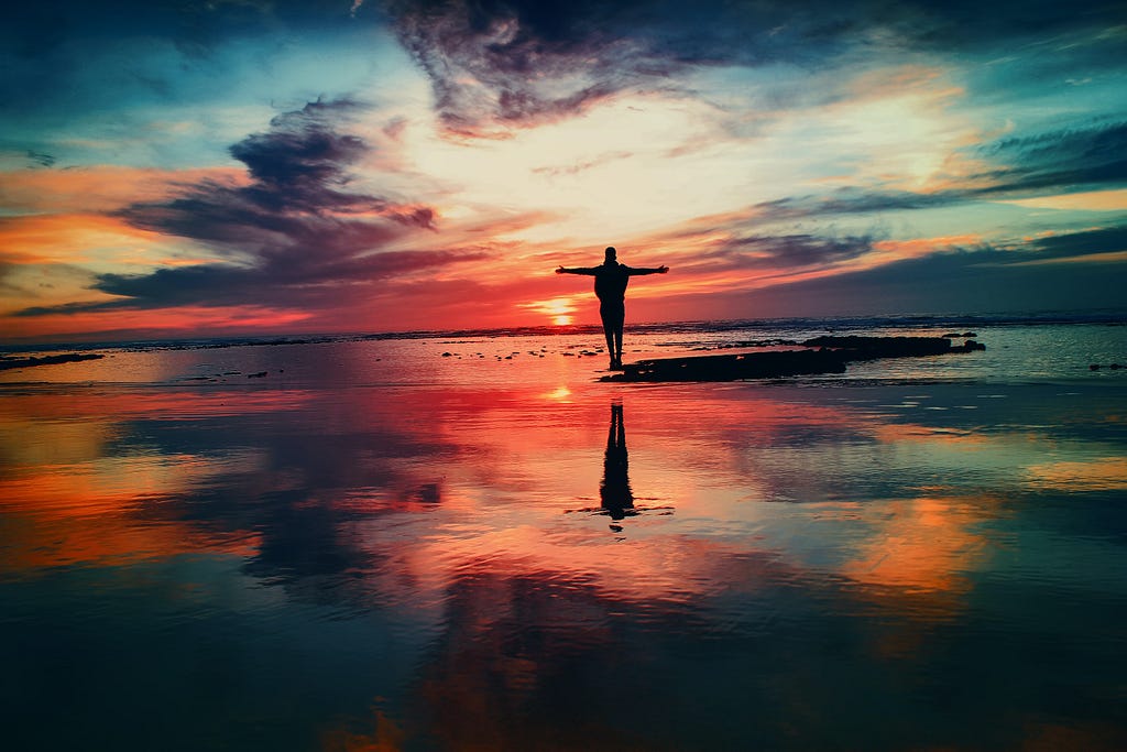 Person silhouetted against the sunset, standing at the edge of wet beach and ocean.