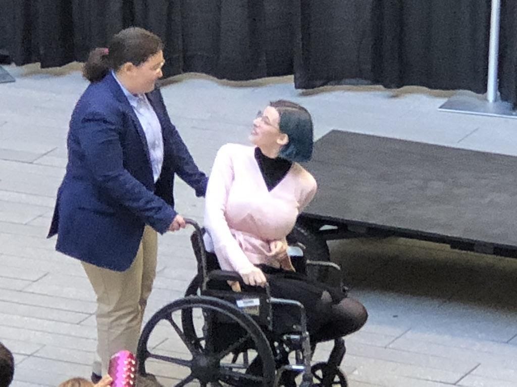 A student actor playing Jessica sits in a wheelchair while the student playing Lorenzo pushes her chair. The students are captured in diologue on stage.