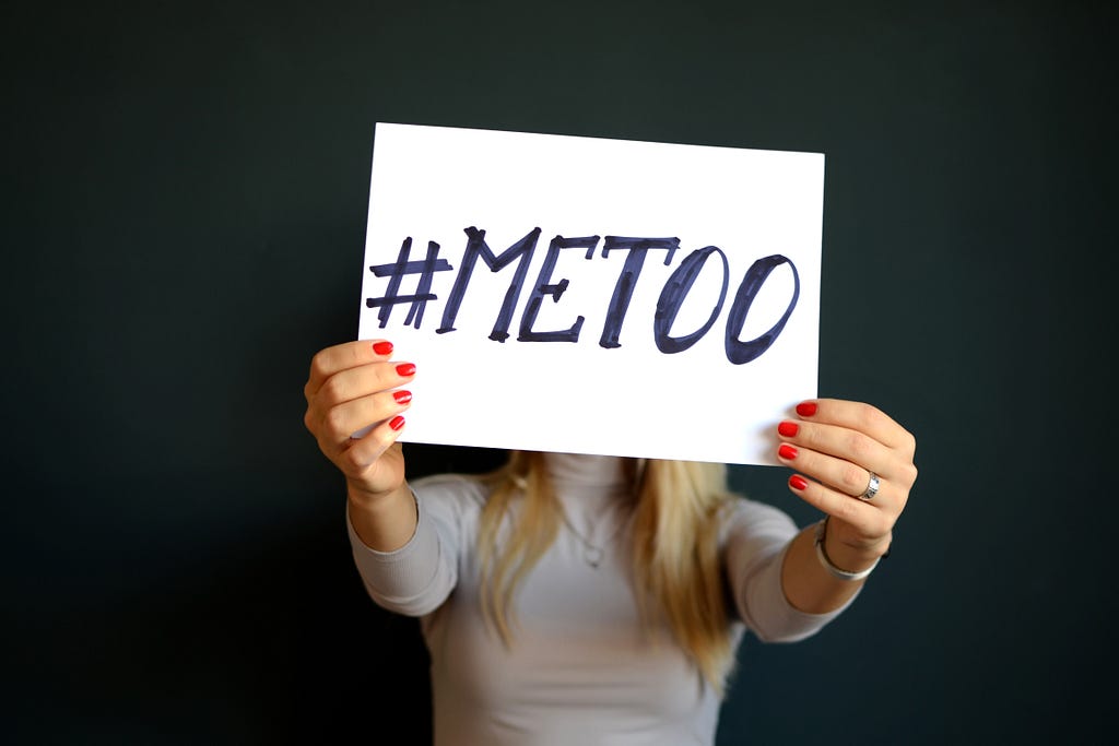 #MeToo escrito en un letrero blanco de tamaño mediano, sostenido por una mujer que vestía una camisa de cuello alto morado.