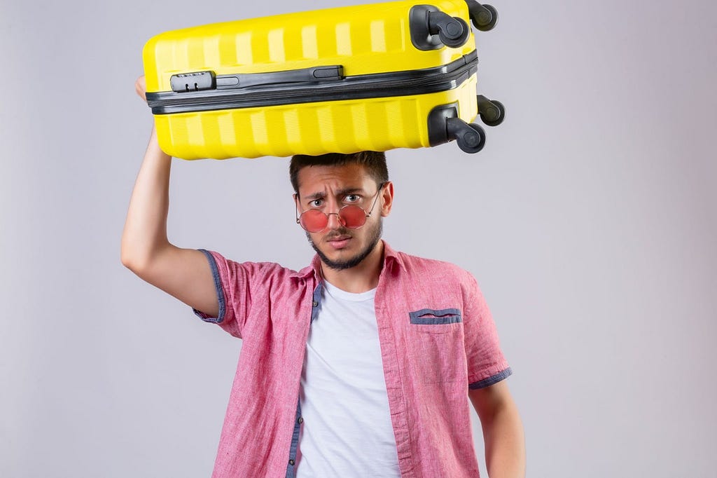 A Latino guy wearing a pink shirt over a white round-neck carries a yellow suitcase on his head