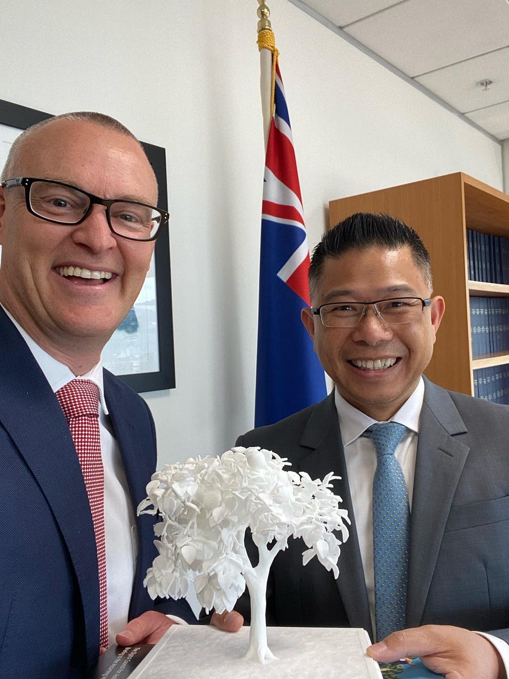 Hon Dr David Clark stands left with Mitchell Pham, right. Between them they hold a small, white 3D printed tree on a stand.