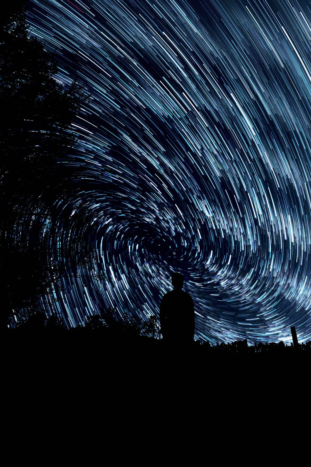 Time-lapse photo of a starry night sky full of cool-coloured stars, generating a swirling vortex of star-light, it’s centre point slightly above a person silhouetted in the foreground.