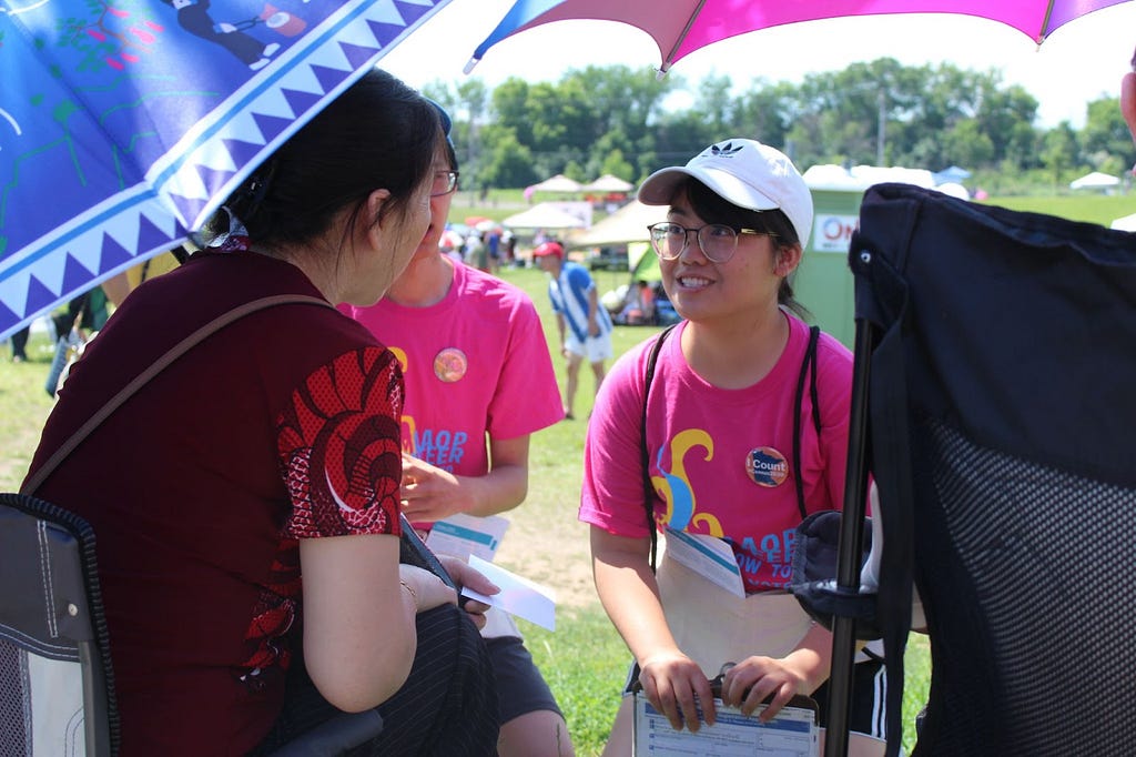 A woman wearing a pink shirt and a white baseball cap speaks with someone at an outdoor event.