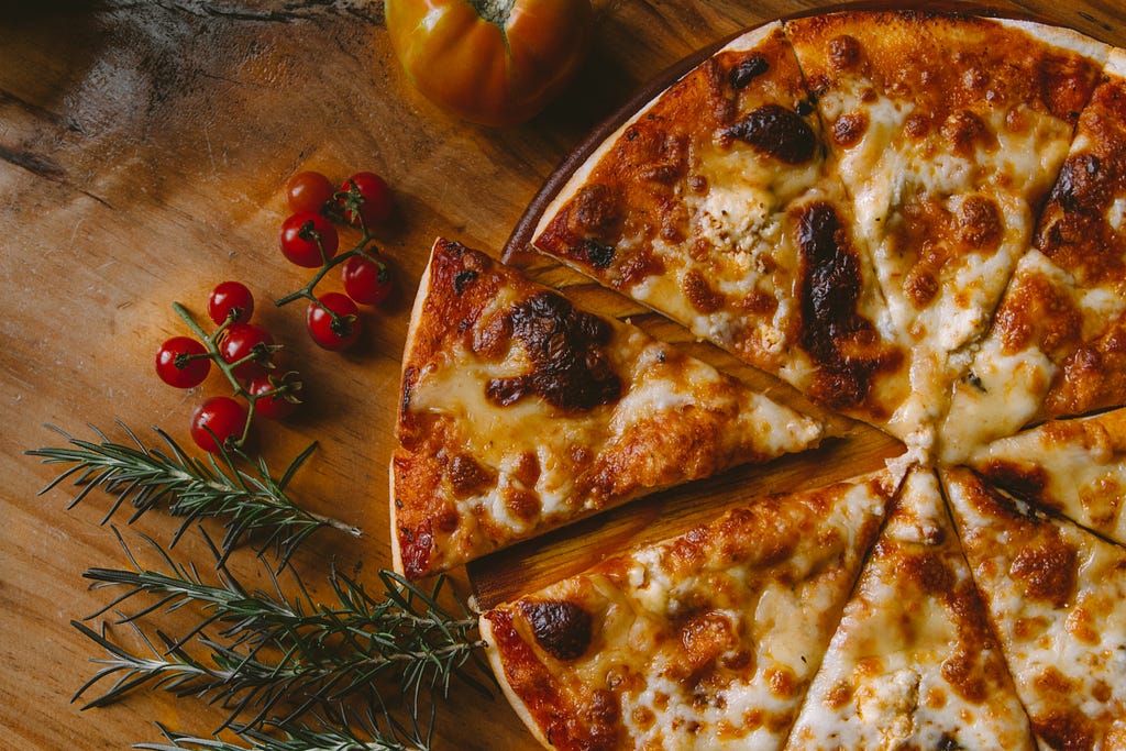 Foto profissional de uma pizza cortada em pedaços, junto com alguns outros ingredientes in-natura como tomate cereja e tomilho.