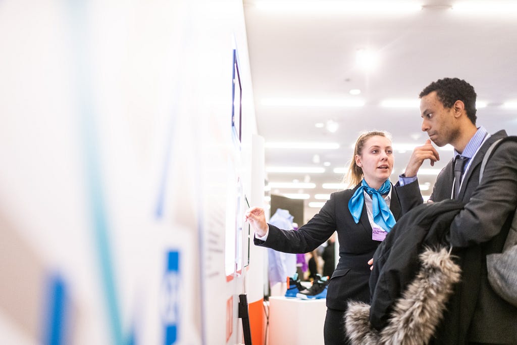 A woman gesturing to a poster presentation, explaining something to a visitor of her stand