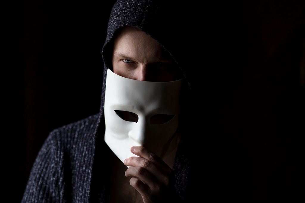An ominous looking man holding a white mask halfway covering his face in a gray hoodie