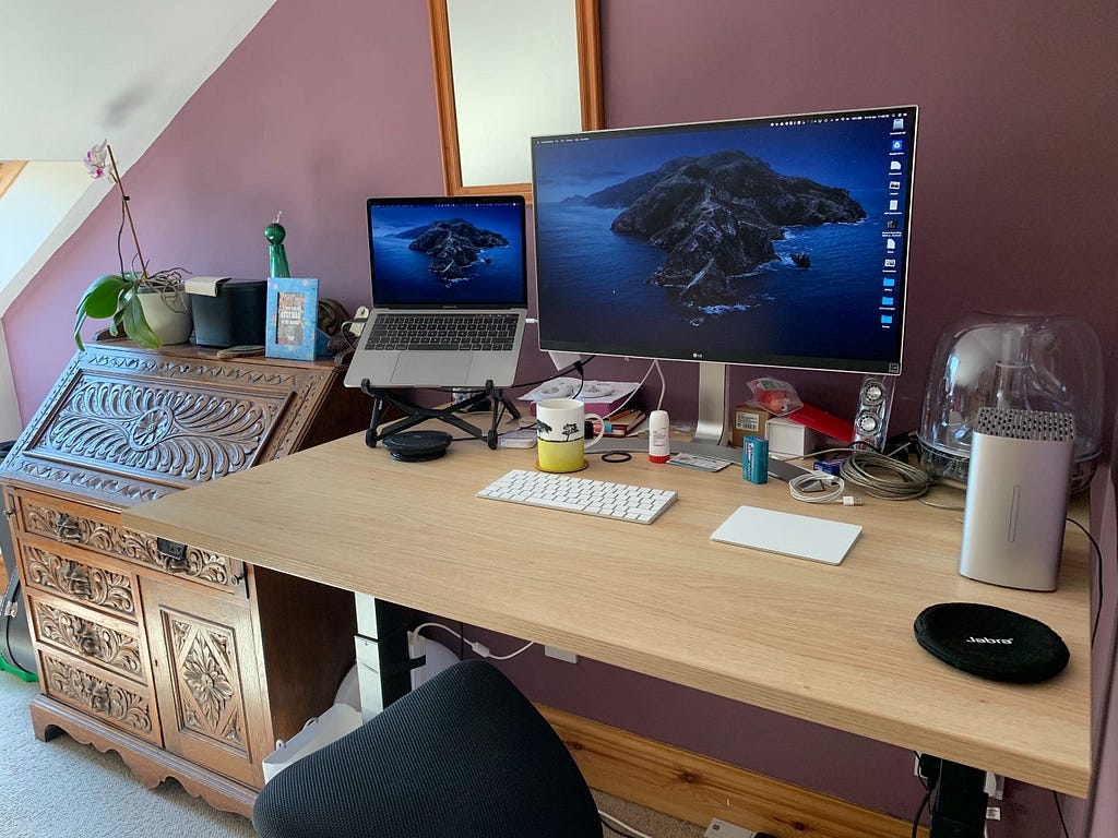 A picture of Joe’s desk in an attic room, with a laptop on a stand, a monitor, speakers, external keyboard and trackpad.