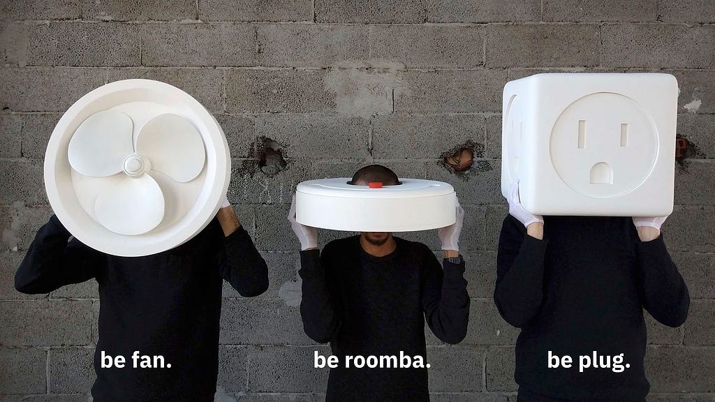 Three people with large household objects on their heads covering their eyes: a fan, a roomba, and a plug.