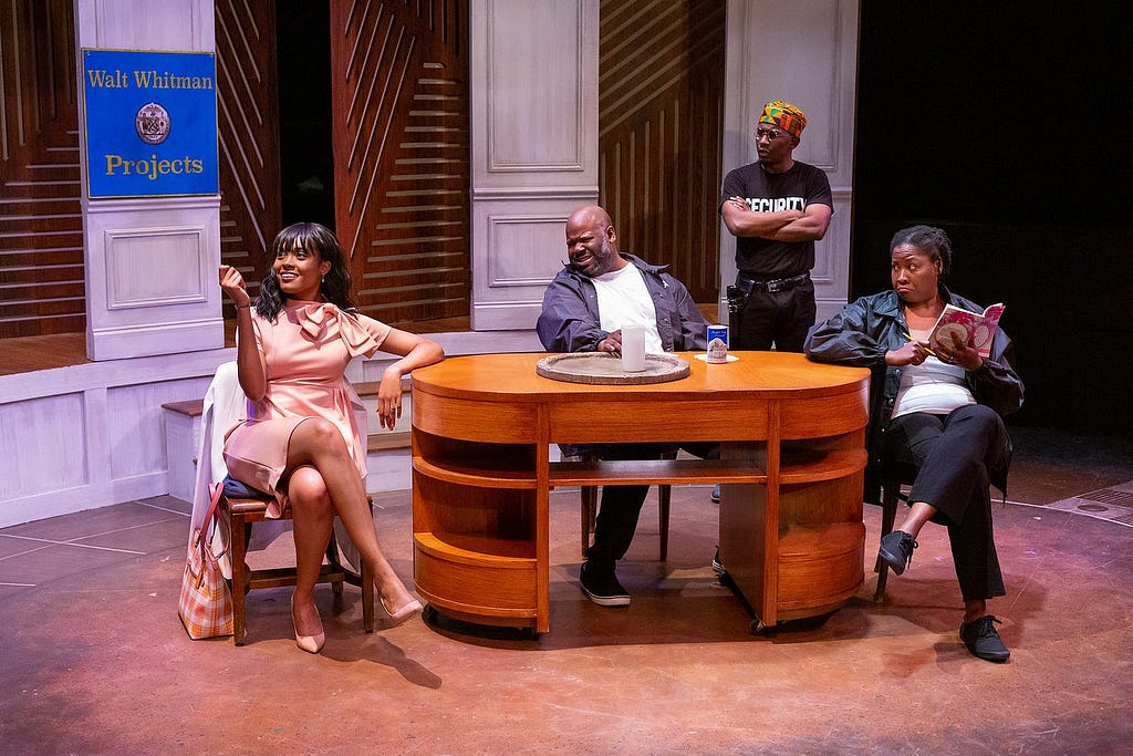 A person in a peach skirt suit sits at a table with three other people, all wearing security guard clothes, onstage.