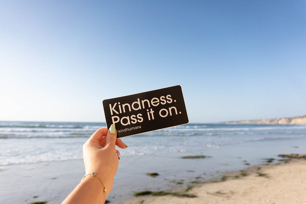 A hand holding a sign on a beach, saying “Kindness. Pass it on.”