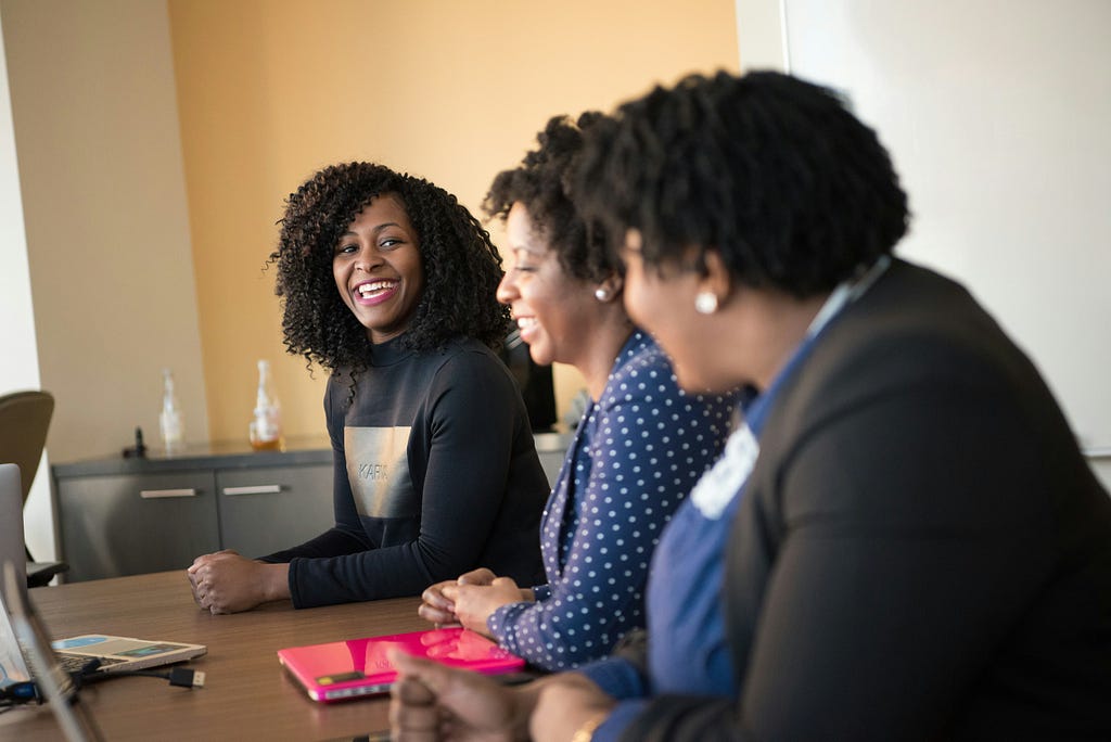 Three women sit together at a conference table engaged in conversation, smiling and laughing during a meeting. Laptops and notebooks are visible on the table, and the atmosphere conveys collaboration, leadership, and teamwork in a professional setting.