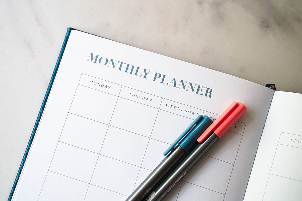 A planner opened to a page that reads “MONTHLY PLANNER” above columns for planning. Two pens, one blue and one pink, lie on the page.