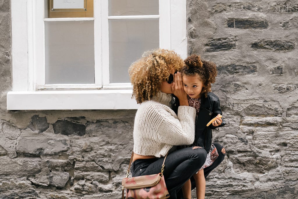 A woman with her daughter, outdoors, in front of a wall. The woman is wispering something in the girl’s ears. She is wearing a white sweater and black trousers.