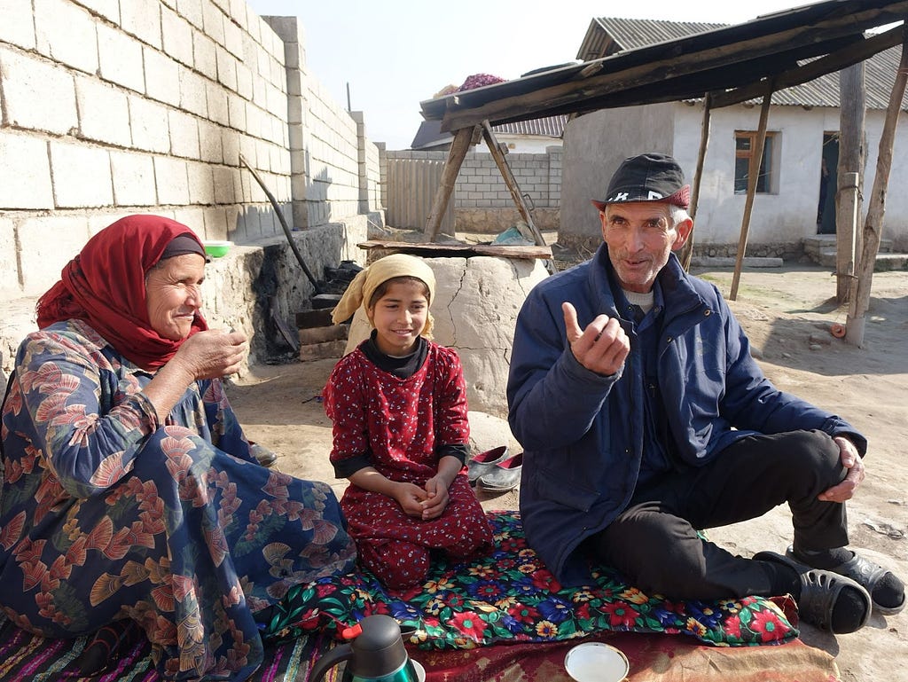 A man gestures while sitting on a colorful blanket with his wife and daughter outside of their home.