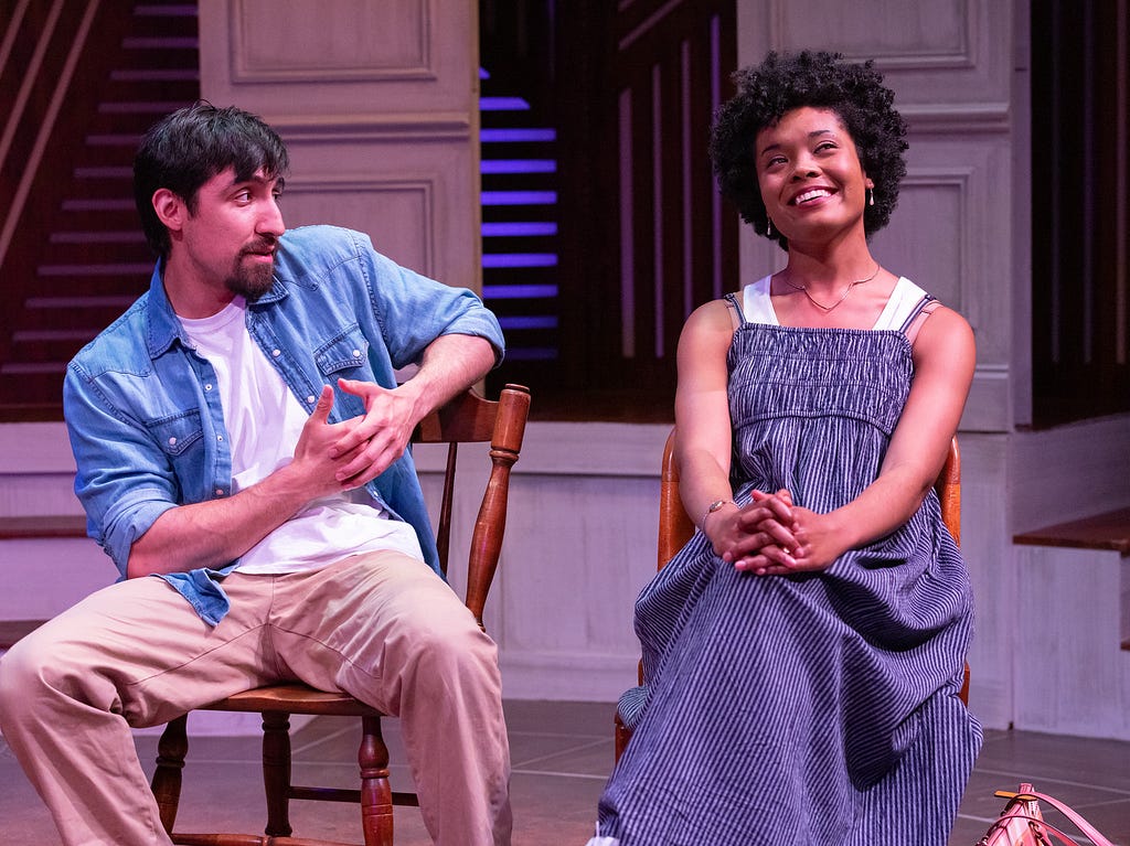 A man in tan pants, a white t-shirt, and an unbuttoned denim shirt with tan skin and dark brown hair and goatee sits looking at the person on the right. She has brown skin and dark brown hair and wears a blue sleeveless dress, and is smiling.