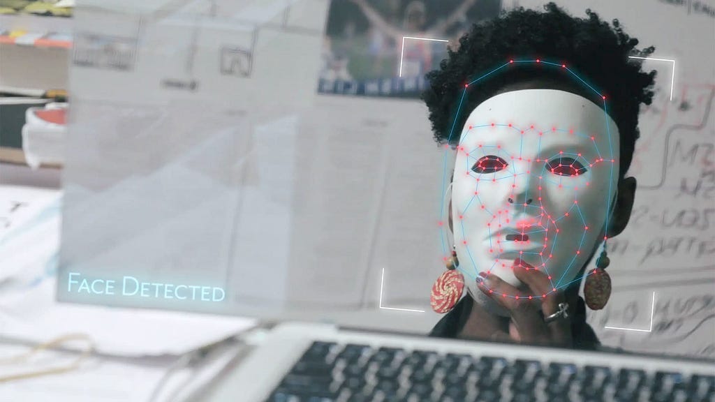 black woman with a white mask being scanned by technology, labeled “face detected”