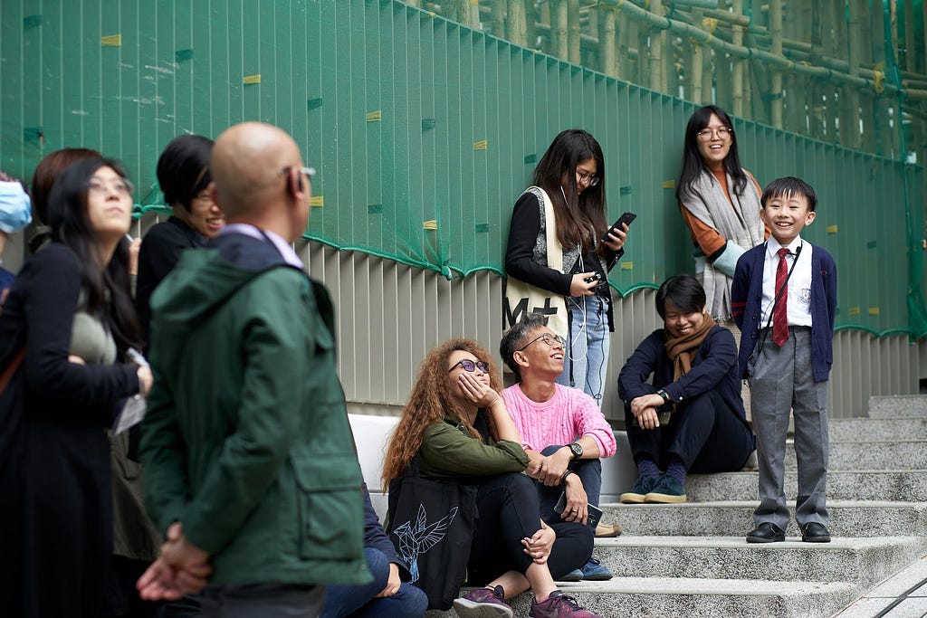 A young student on a staircase speaks to a smiling group of people around him, surrounded by bamboo installation artwork.