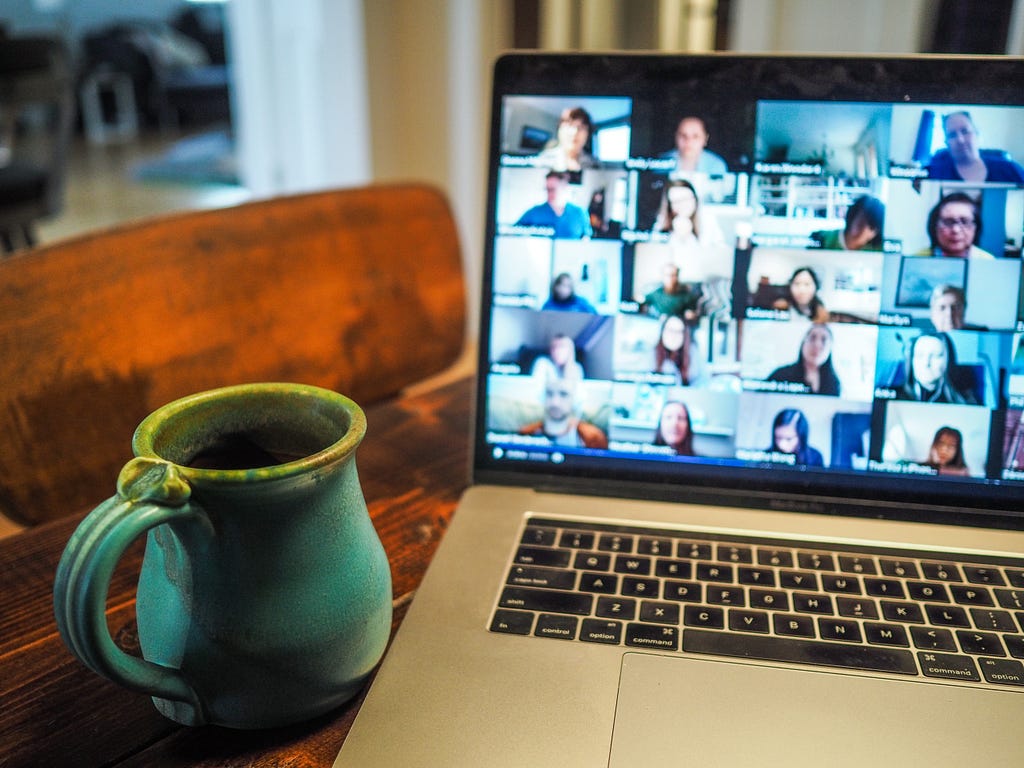 A laptop and a mug of coffee on a table. The laptop shows images of 20 people who are on a video conference call.