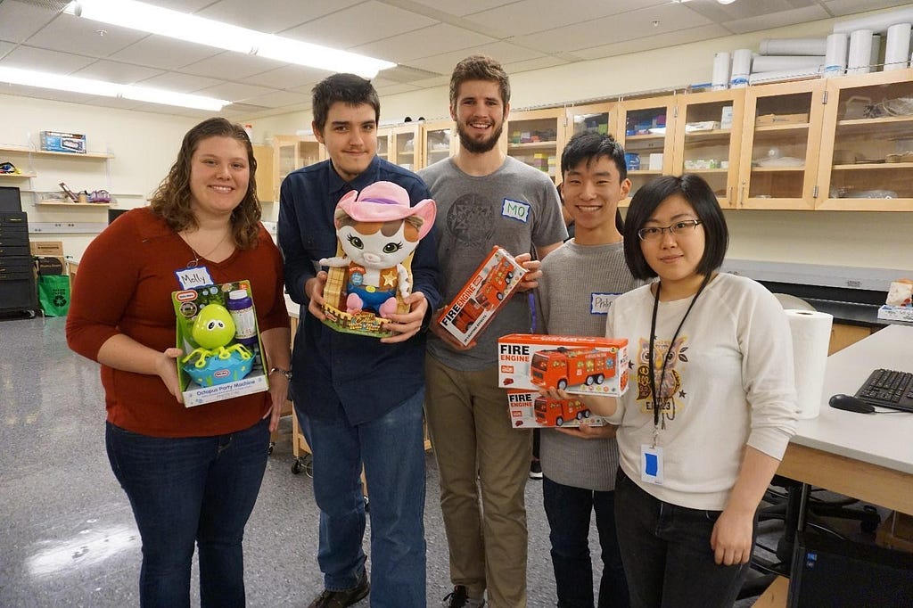 Four people hold boxes with toys. In the background there are workbenches and drawers.