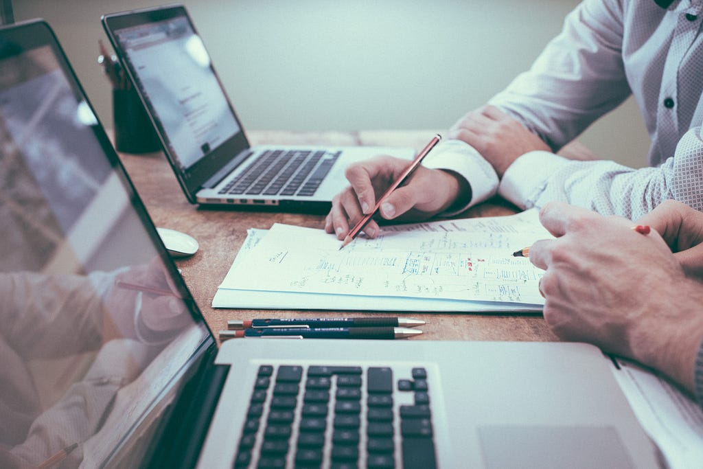 A person explains, with the help of notes, something to another person. Both sit in front of their laptop. They wear workplace attire. Only the hands are visible.