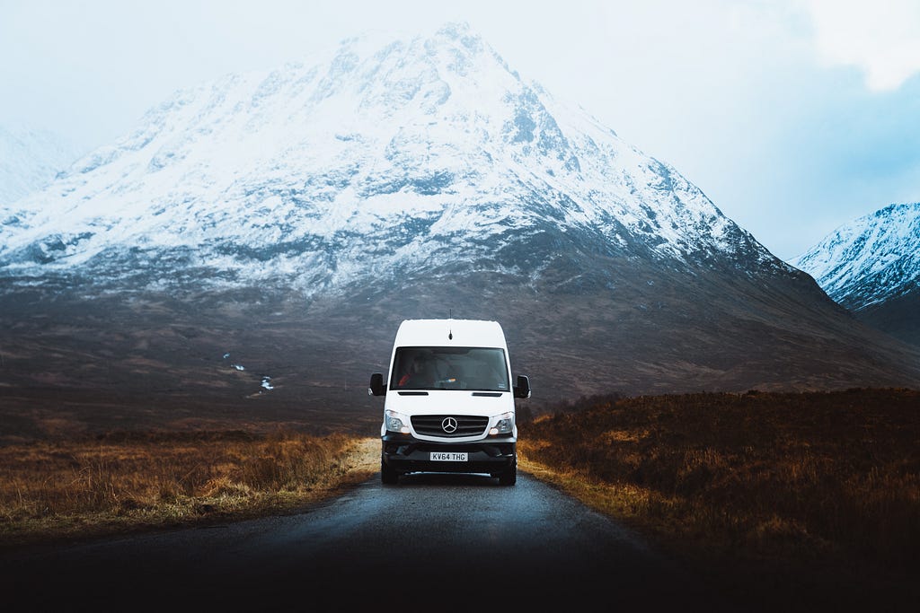 A high roof sprinter van driving down a road with snowy mountains behind it.